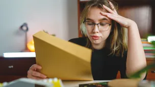 Jente eller ung kvinne med blondt og hår og briller som ser stresset ut og holder papir i den ene hånda