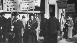 Åtte kvinner går langs fortauet med plakater på det som trolig er Norges første 8. mars markering. Parolene handler om likelønn, fred og nei til atomvåpen