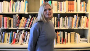 Ung kvinne med lyst, langt hår i blå genser, stående foran en bokhylle i Kvinnemuseet.