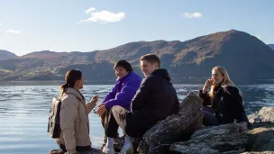 En gruppe unge mennesker sitter ved vannet med fjell i bakgrunnen