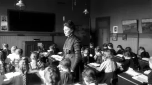 Sort-hvitt-foto av Anna Rogstad stående i klasserommet mens barna sitter bøyd over pultene sine i 1909