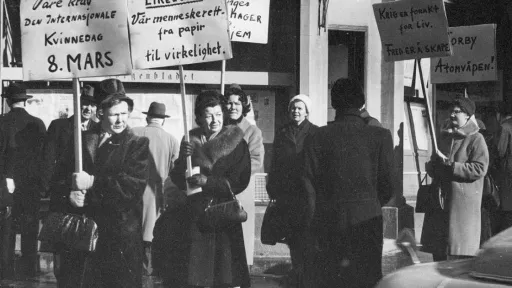 Åtte kvinner går langs fortauet med plakater på det som trolig er Norges første 8. mars markering. Parolene handler om likelønn, fred og nei til atomvåpen