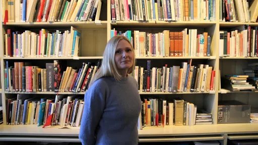 Eva Refsahl, ung kvinne med lyst, langt hår i blå genser, stående foran en bokhylle i Kvinnemuseet.