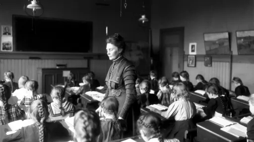Sort-hvitt-foto av Anna Rogstad stående i klasserommet mens barna sitter bøyd over pultene sine i 1909