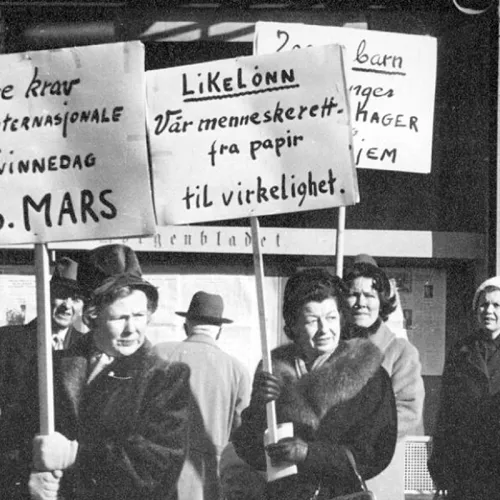 Svart-hvitt bilde av to kvinner i mørke kåper. De holder hver sin plakat med teksten "Våre krav den internasjonale kvinnedag 8. mars", og "Likelønn, vår menneskerett -fra papir til virkelighet".