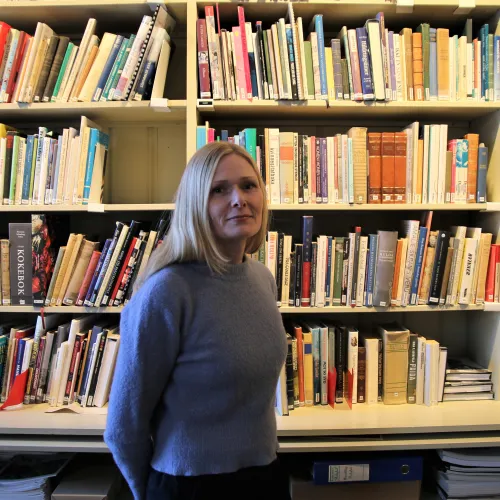 Eva Refsahl, ung kvinne med lyst, langt hår i blå genser, stående foran en bokhylle i Kvinnemuseet.