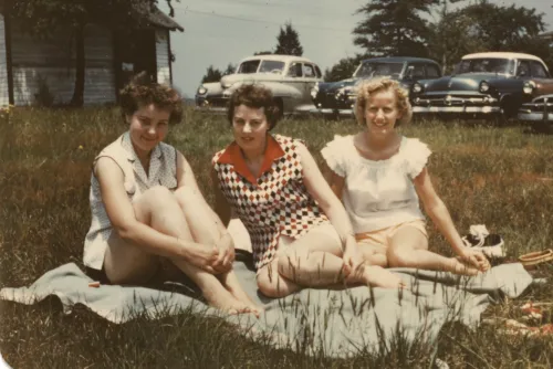 Tre kvinner fotografert i New Jersey på 1950-tallet, i sommerklær sittende på et teppe på en gresslette med amerikanske biler parkert i bakgrunnen